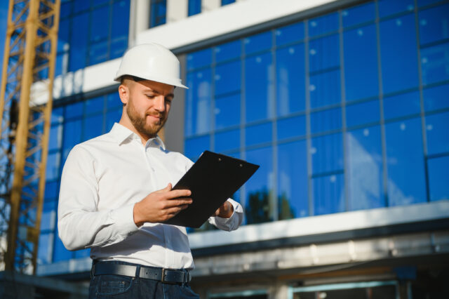 Engineer Next To Building Cranes. Concept Large Construction Project. Architect Directs The Construction Process. Drawings And Tablet In The Hands.