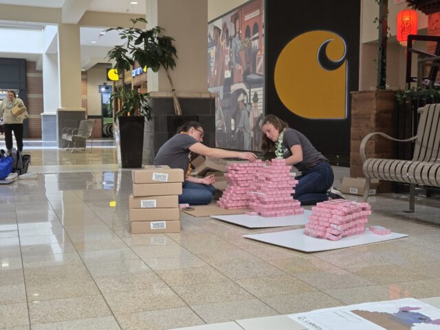 Two people build a structure out of boxes of food