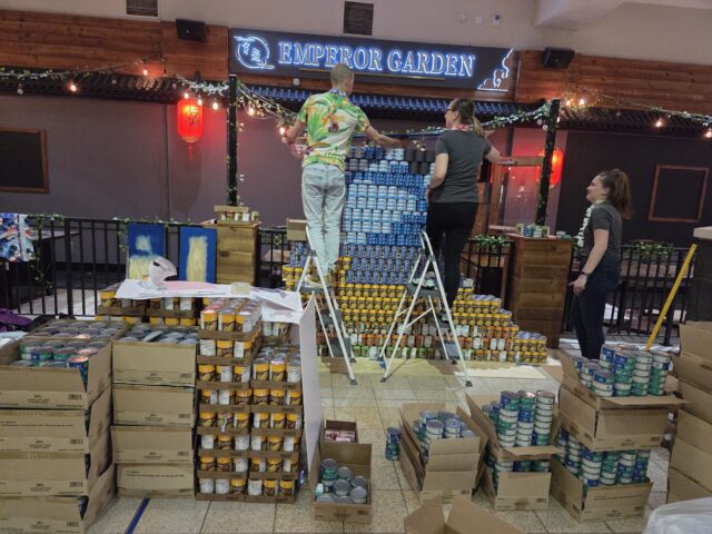 Three people, two on ladders, stack cans to build a structure.