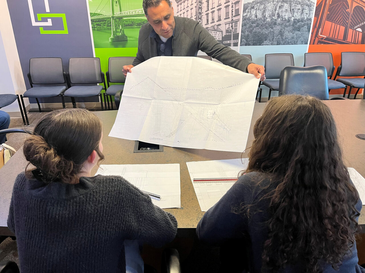 A photo of a person holding an architectural plan up for two younger individuals to see, in an office setting.