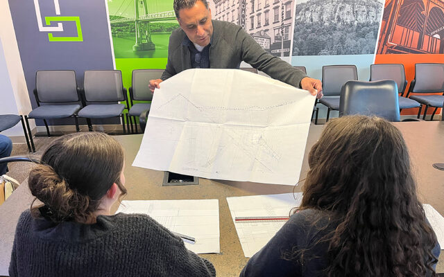 A photo of a person holding an architectural plan up for two younger individuals to see, in an office setting.